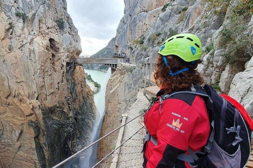 A person in a red jacket and green helmet standing on a bridge overlooking a canyon with a waterfall. 1