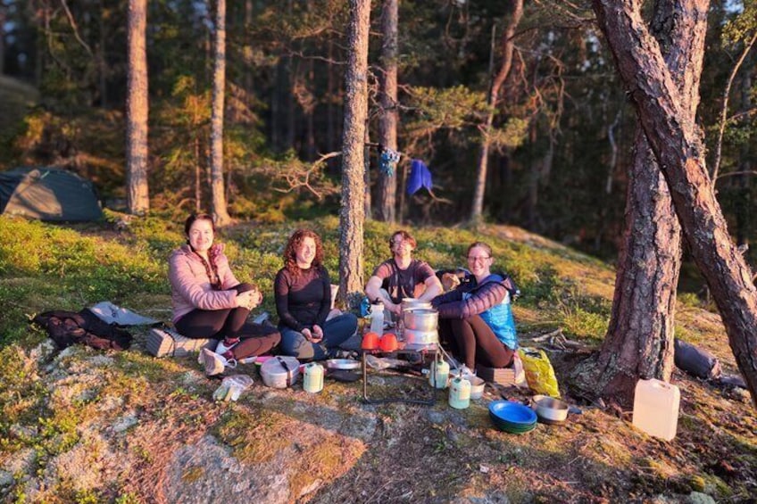 Cuatro personas sentadas en el suelo de un bosque, con equipo de campamento y una olla sobre un hornillo portátil. 3