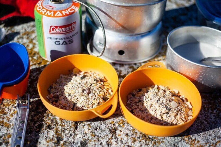 Dos tazones amarillos con comida, un bidón de gas Coleman y utensilios de cocina para camping sobre una superficie rocosa. 4