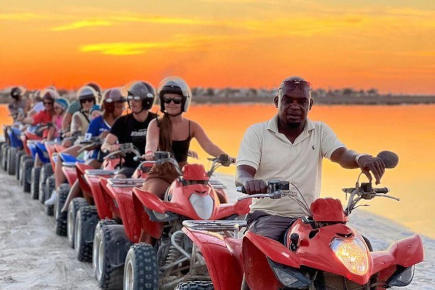 Un groupe de personnes à bord de quads s’est aligné sur un chemin de sable, au bord d’une étendue d’eau, au moment du coucher du soleil. 1