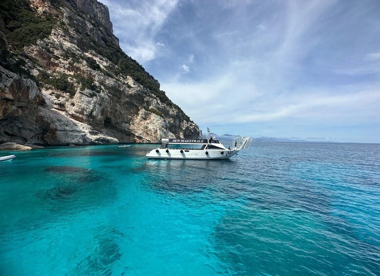 A boat named 'MIN MAESTRALE' anchored near a rocky coastline with clear turquoise waters. 1