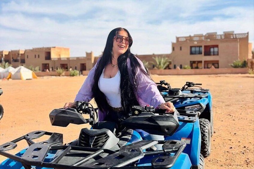 Une personne est assise sur un quad bleu dans un désert, avec des tentes et des bâtiments en arrière-plan. 1
