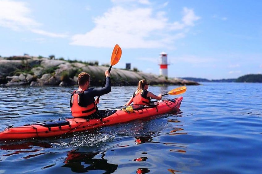 Two people kayaking on a calm body of water with a lighthouse in the background. 1