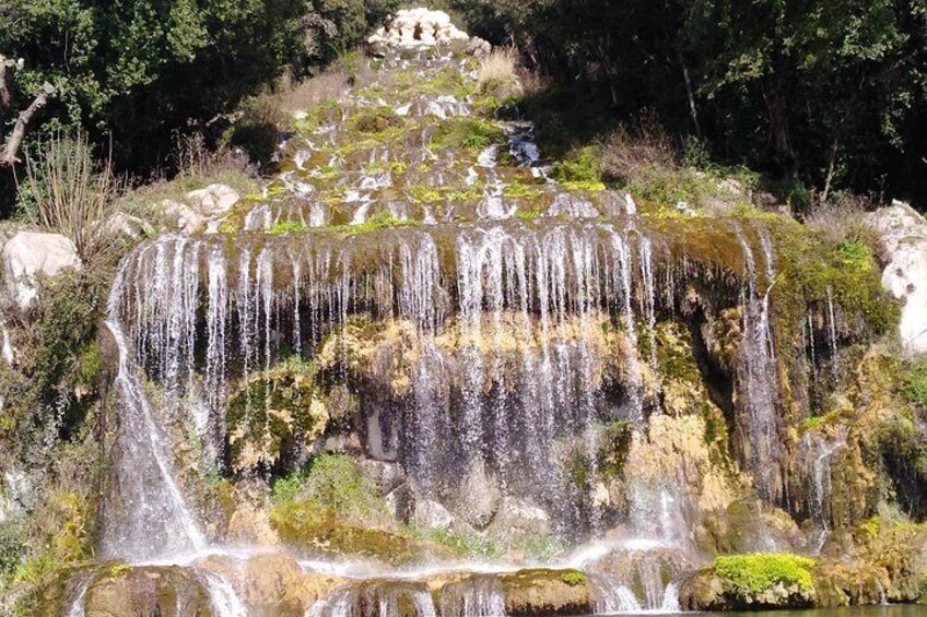 Ein mehrstufiger Wasserfall stürzt über moosbewachsene Felsen, umgeben von dichtem Grün. 3