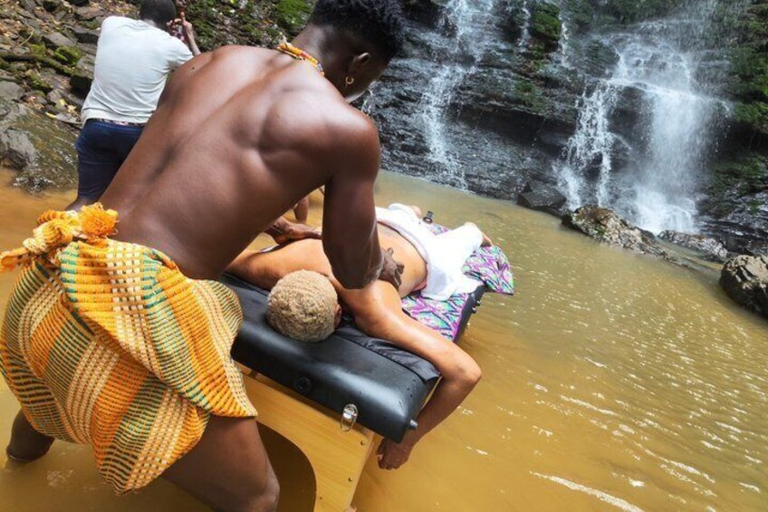 A person in traditional attire assisting another person on a wooden platform near a waterfall. 2