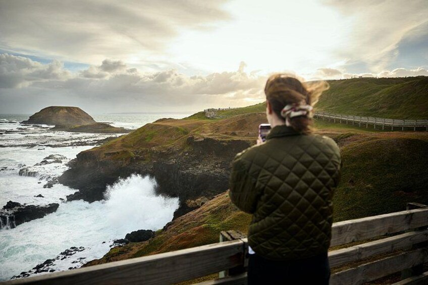 A person taking a photo of a coastal landscape with cliffs and a bridge. 4