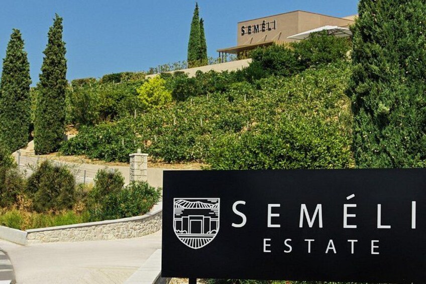 Skilt for Semeli Estate med logo, omgivet af vegetation og træer. 3