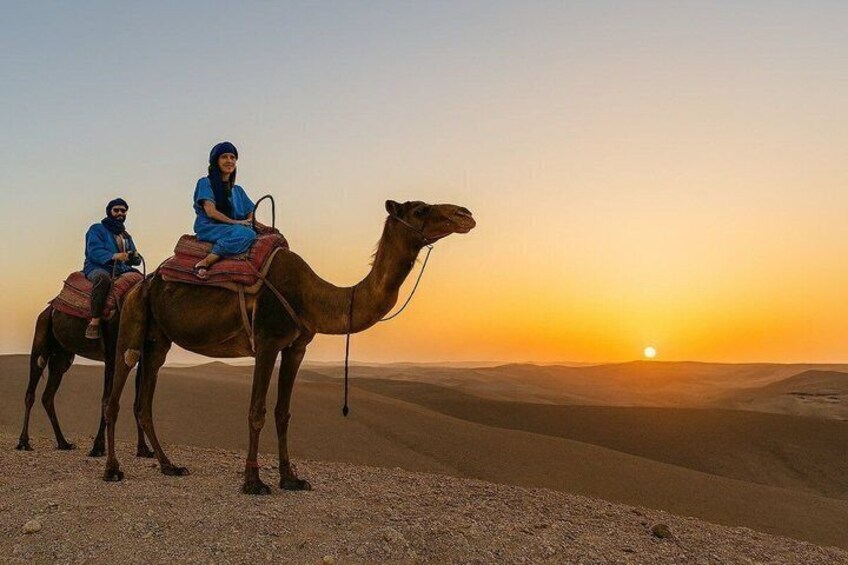 Dos personas montando un camello en un desierto, con un atardecer al fondo. 3