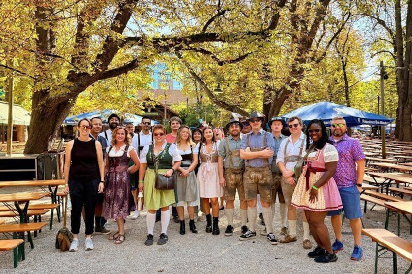 A group of people outdoors, some in traditional attire, standing under trees with picnic tables around. 5