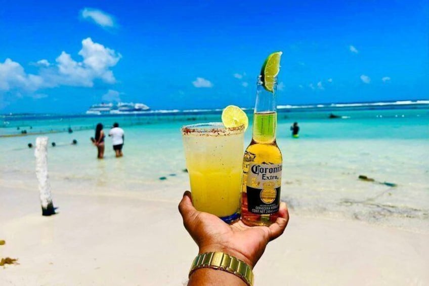 Una mano sosteniendo una margarita y una botella de Corona Extra en una playa, con un crucero al fondo. 1