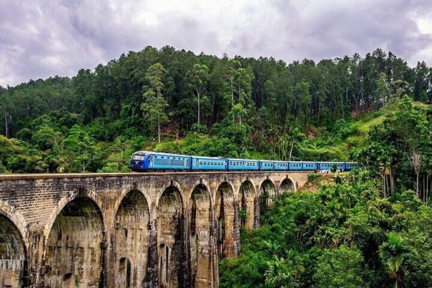 A blue train traveling over an old stone bridge surrounded by lush green forest. 1
