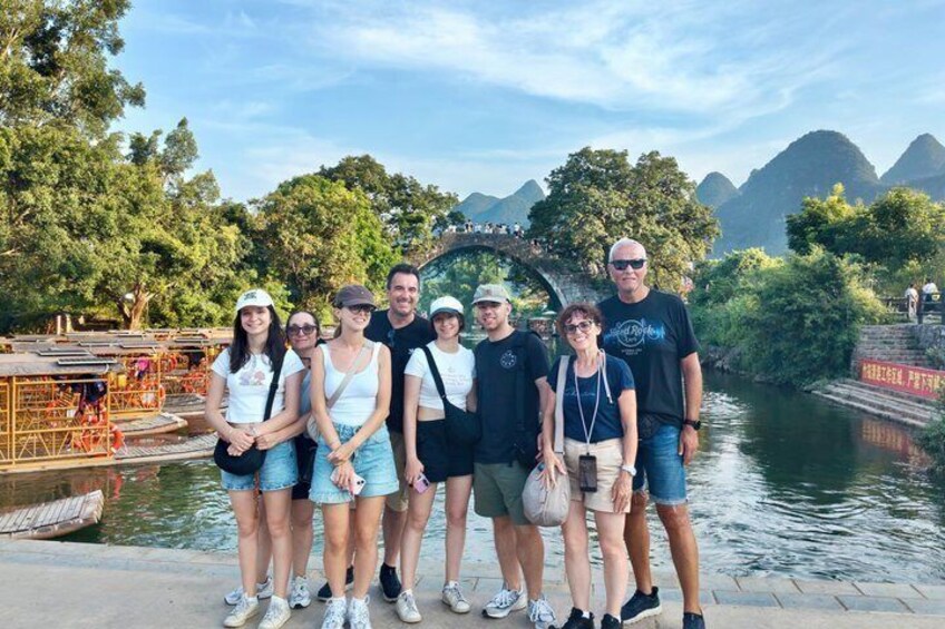 A group of people standing by a river with a bridge and mountains in the background. 2