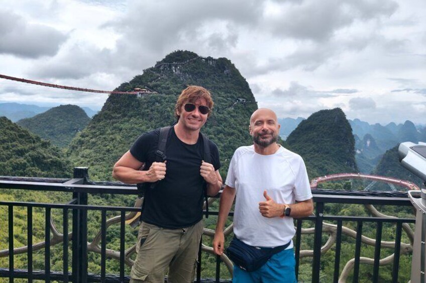 Two people standing on a viewing platform with a mountainous landscape in the background. 3