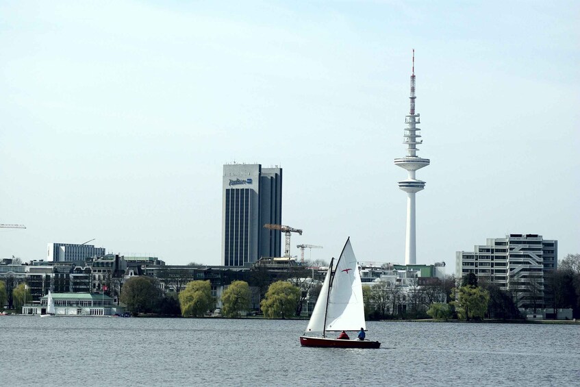 A sailboat on a river with a city skyline in the background, including a tall tower. 3