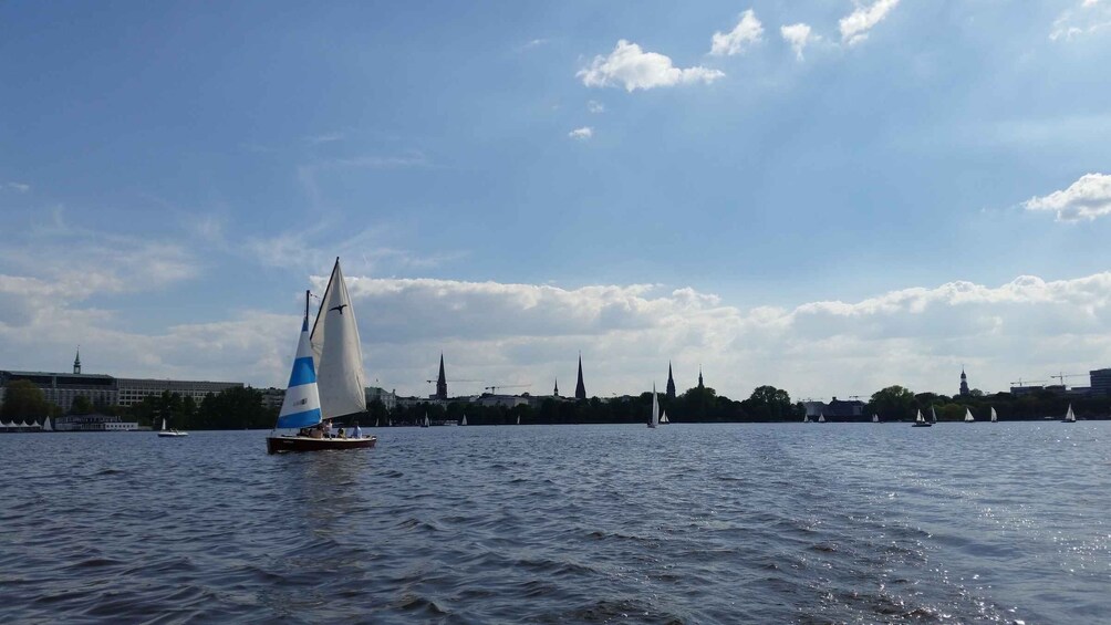 A sailboat with a blue and white sail on a river, with a city skyline in the background. 2