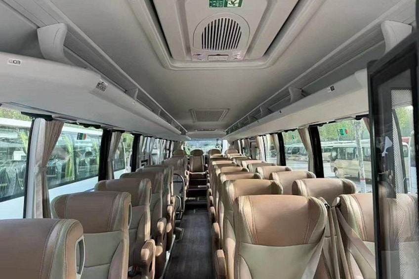 A bus interior with rows of beige seats, overhead storage compartments, and a central aisle. 2