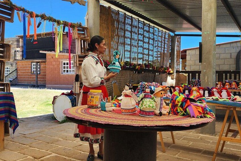 A person standing at a table with colorful dolls and textiles, with a building and a clear sky in the background. 2