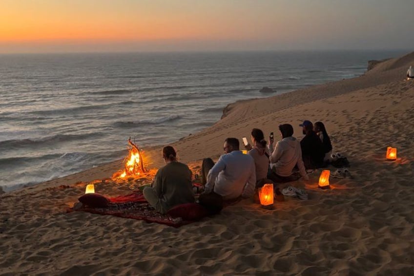 A group of people sitting on a beach at sunset, with a fire in the center and lanterns around. 1