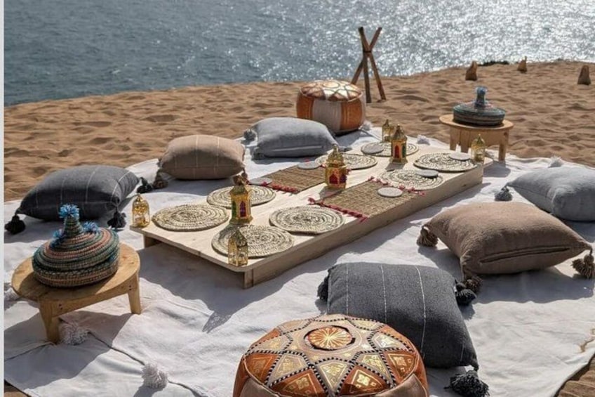 A beach setting with a picnic setup including a blanket, pillows, woven mats, and decorative items. 3