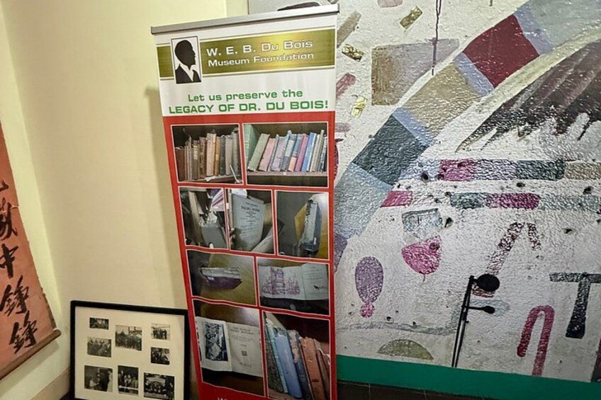Une bibliothèque remplie de livres et de photos encadrées, un mur orné d’une mosaïque et une affiche faisant la promotion de la Fondation du musée W.E.B. Du Bois. 5