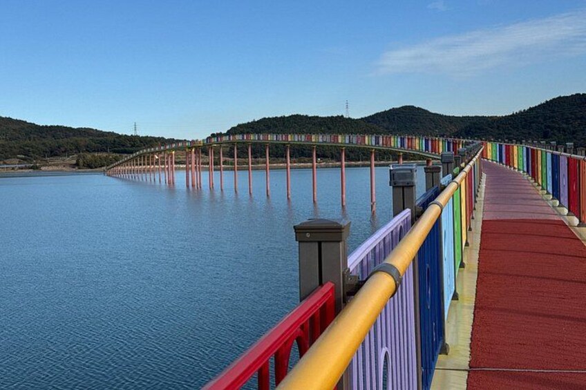 A long, colorful pedestrian bridge over a body of water with a red walkway and a yellow railing. 1