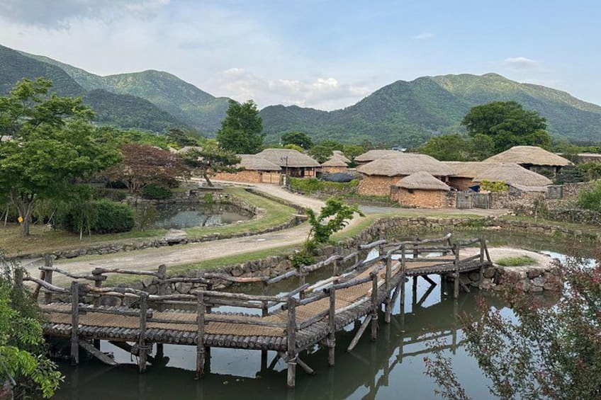 A traditional village with thatched-roof houses, a wooden bridge over a pond, and mountains in the background. 2
