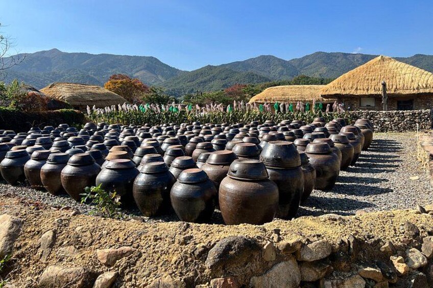 A collection of large pottery pots arranged in rows, with a thatched-roof building and mountains in the background. 3