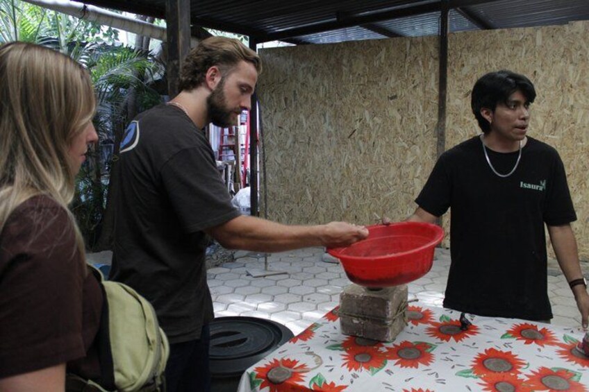 A person pouring water from a red basin into a container on a table with a floral tablecloth. 5