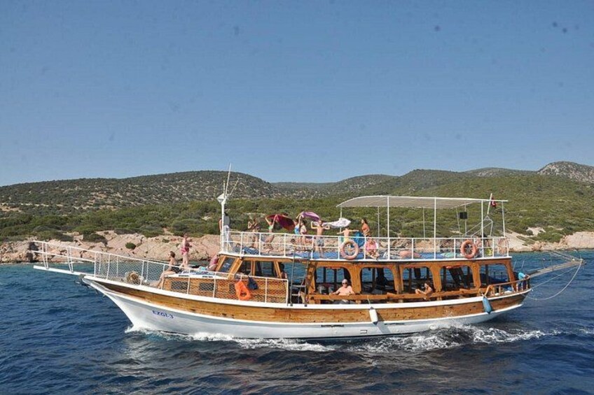 A wooden boat with passengers, moving on calm water with a rocky coastline and hills in the background. 1