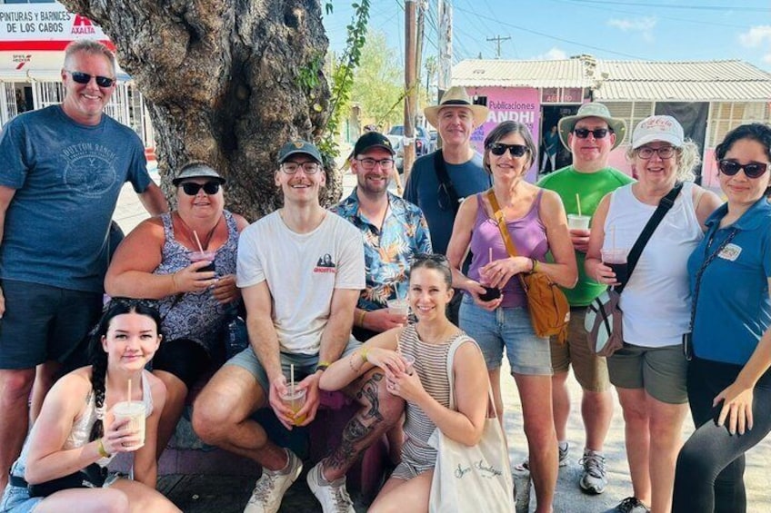 En grupp människor som poserar utomhus, några håller i drinkar, med ett träd och byggnader i bakgrunden. 5