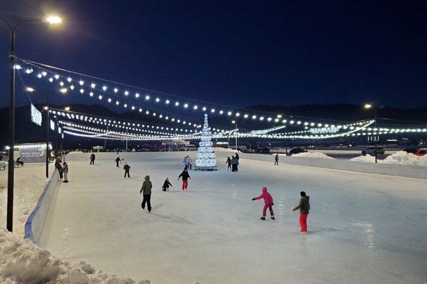 An ice skating rink at night with people skating, a large illuminated Christmas tree, and string lights overhead. 3