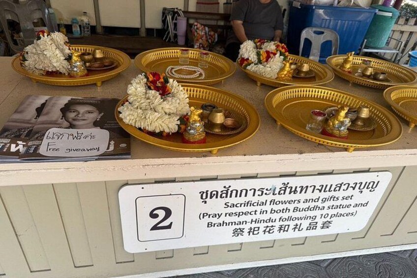 A table with offerings of flowers and gifts, a sign with Thai text, and a numbered placard. 2