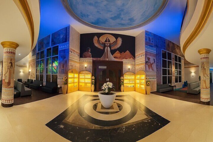 Eine Lobby mit einem Wandgemälde einer ägyptischen Figur, einer großen Pflanze im Topf und einem Empfangstresen. 1