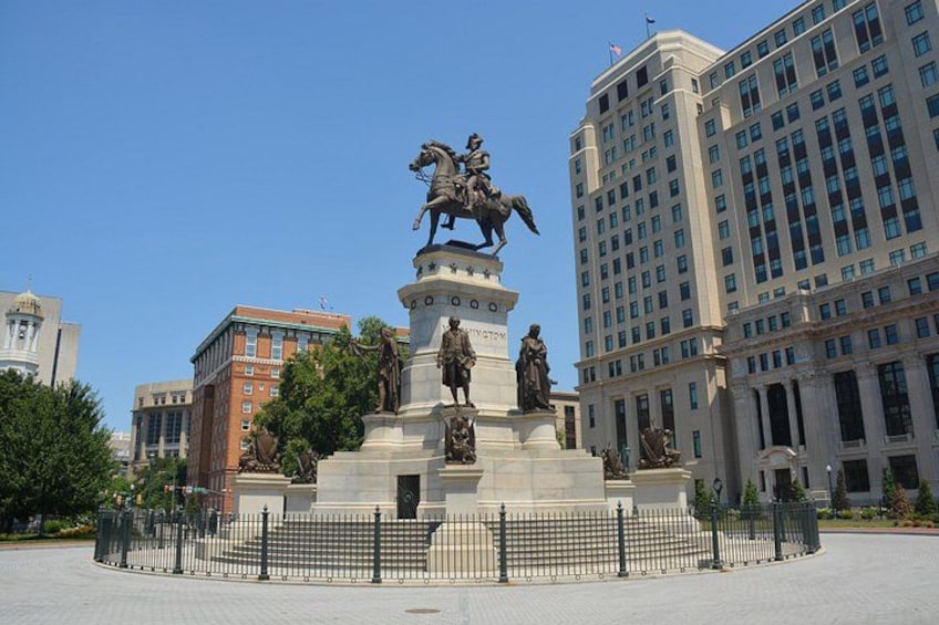 Ein historisches Denkmal mit Statuen, umgeben von einem Zaun, in einer städtischen Umgebung mit Gebäuden. 1