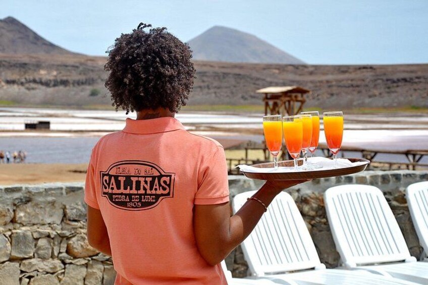 A person in a pink shirt holding a tray with four glasses of orange juice, overlooking a beach with mountains in the background. 4