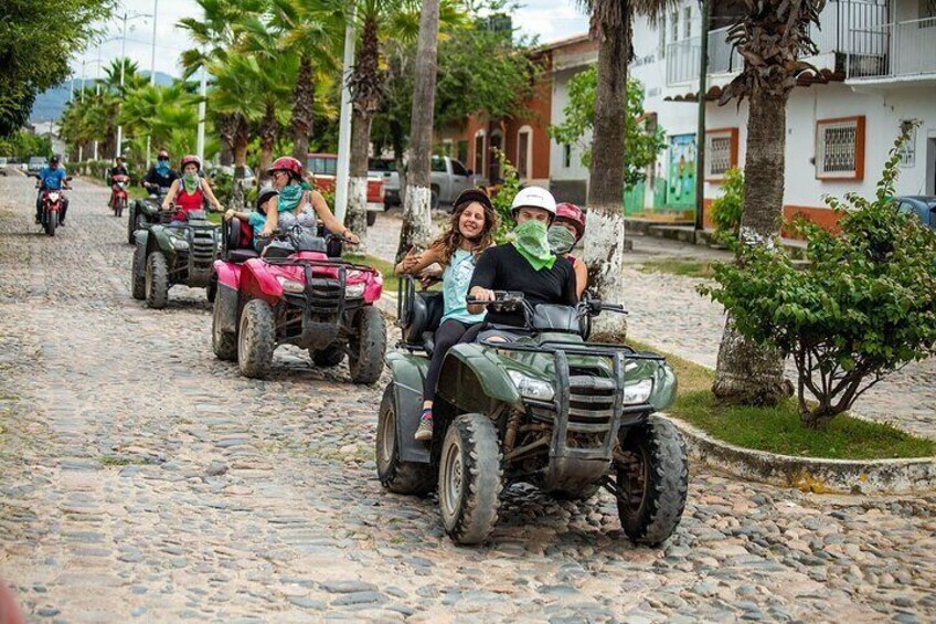 People riding ATVs on a cobblestone street lined with palm trees and buildings. 2