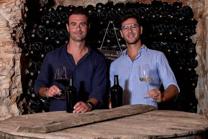 Two men holding wine glasses in front of a wine cellar with bottles. 1
