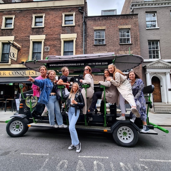 A group of people on a green and black three-wheeled vehicle with a canopy, parked on a street with brick buildings and a sign reading 'The Conquest'. 2