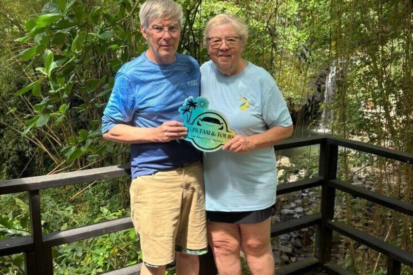 Dos personas de pie sobre un puente de madera, con follaje y una cascada al fondo. 4
