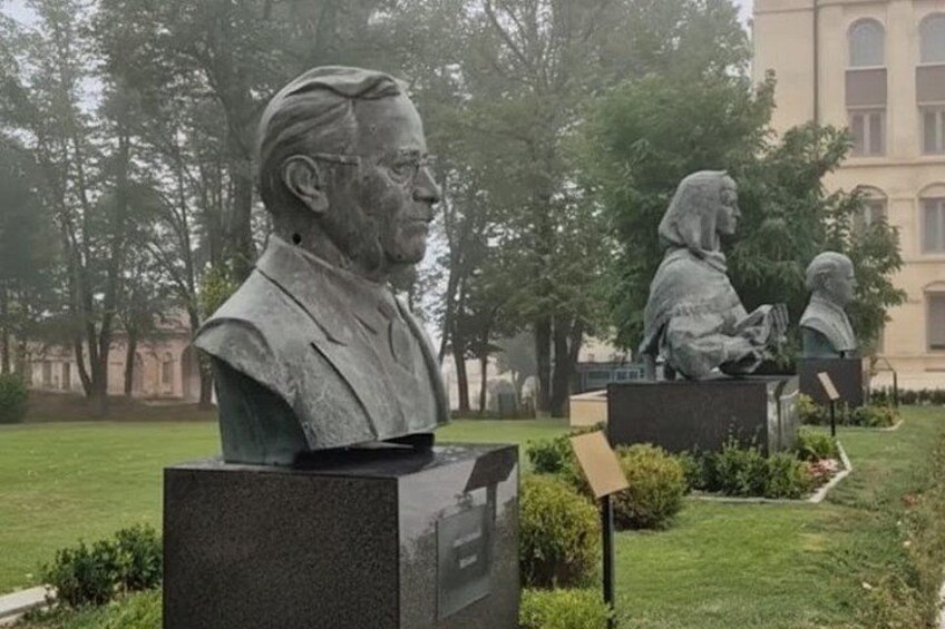 Una estatua de un hombre trajeado y una mujer leyendo un libro en un parque con árboles y un edificio al fondo. 2
