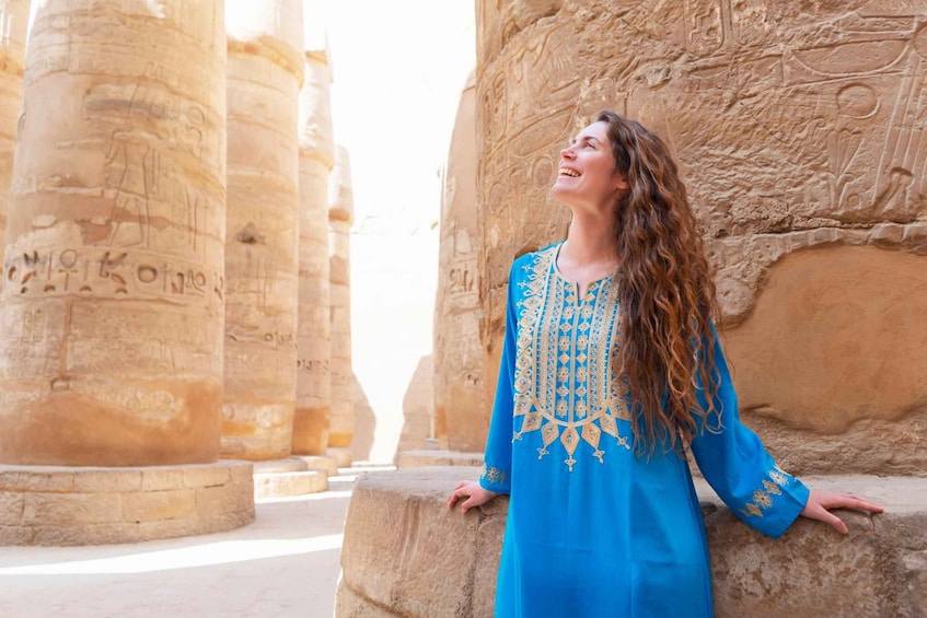 A person in a blue dress with gold embroidery standing in front of ancient stone columns with hieroglyphs. 4