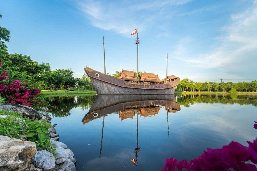 En traditionel båd med flag, fortøjet i roligt vand, omgivet af grønt og blomster. 2