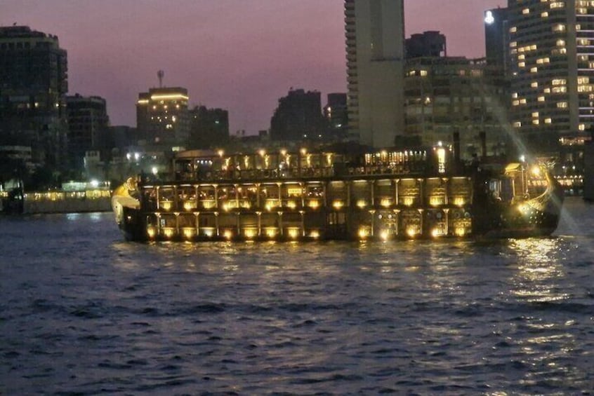 A brightly lit riverboat with multiple decks, surrounded by city buildings at dusk. 2