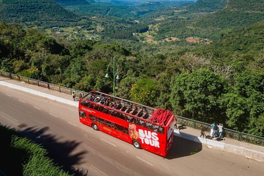 A red double-decker bus labeled 'BUS TOUR' on a road with a scenic view of green hills and trees. 1