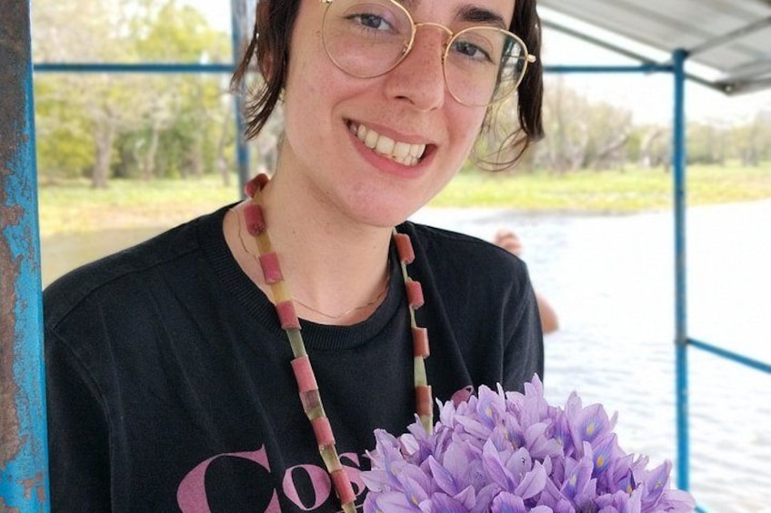 A person holding a purple flower, wearing a black shirt with a visible logo, and standing under a blue structure. 4