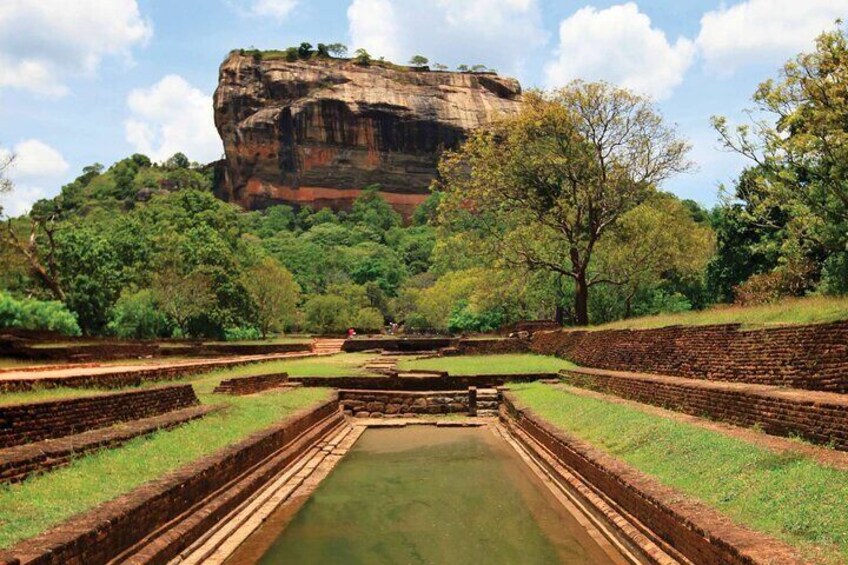 A historical site with a large rock formation, terraced structures, and a central water feature. 2