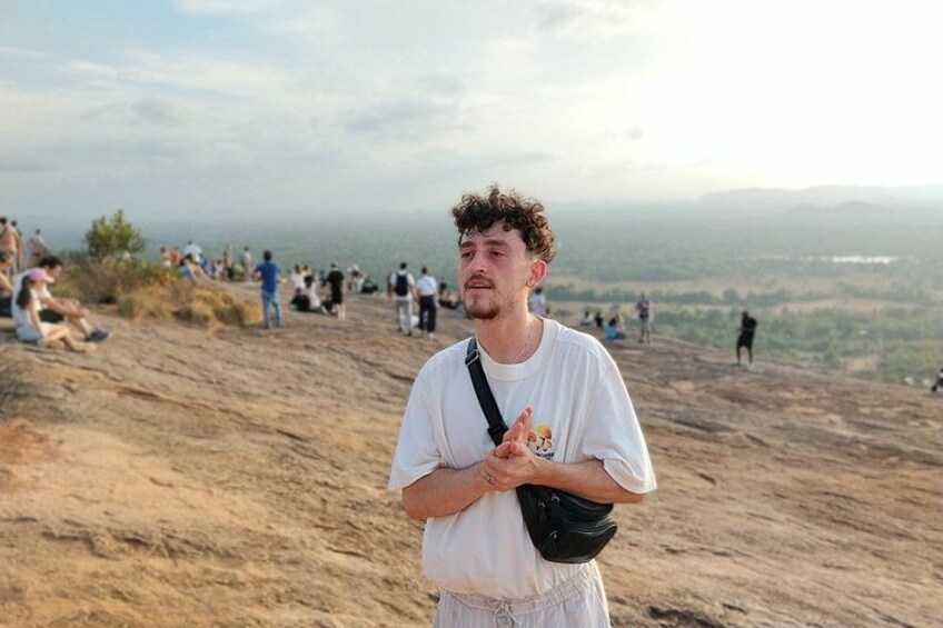 A person standing on a hill with a bag, overlooking a crowded beach and distant landscape. 1