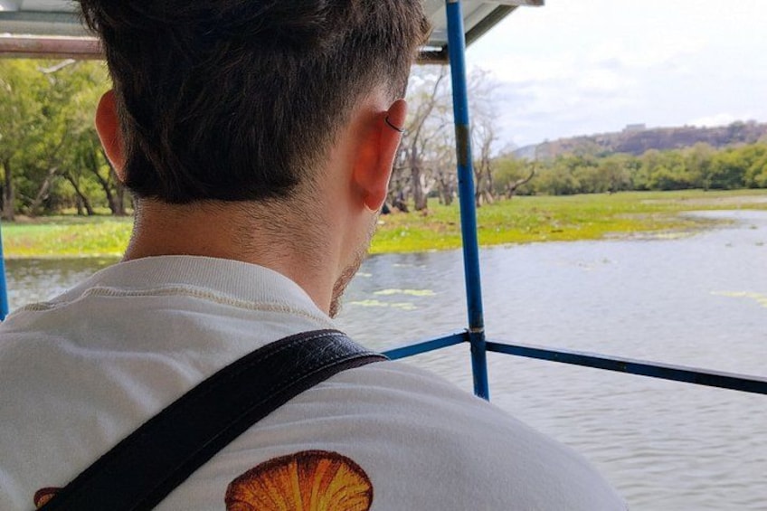A person wearing a white shirt with a shell logo, looking out over a body of water with greenery and trees in the background. 3