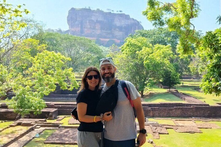 Two people standing in front of ancient ruins with a large rock formation in the background. 1