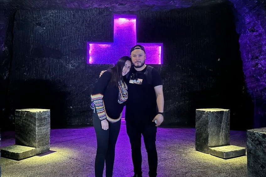 Two people standing in a cave with a large purple cross on the wall. 4